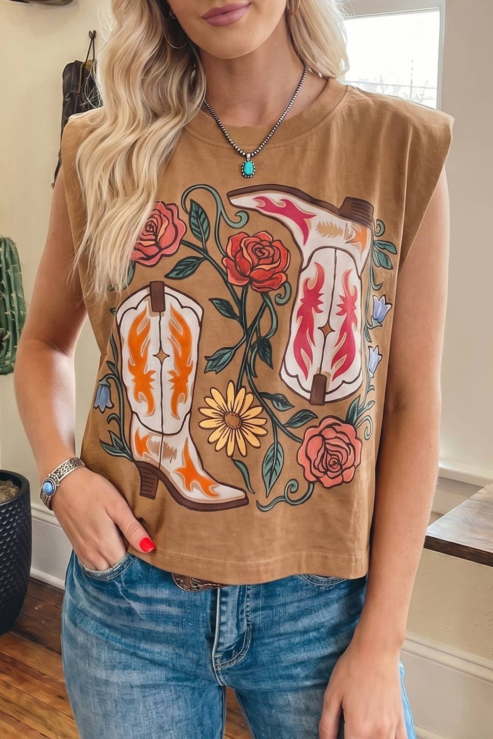 Woman wearing a sleeveless brown top with floral and abstract designs, paired with blue jeans indoors.