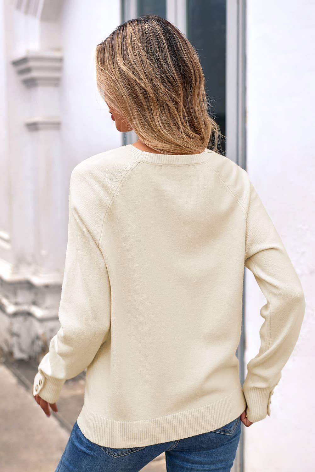 Woman wearing a cream sweater and blue jeans, standing in front of a glass door.