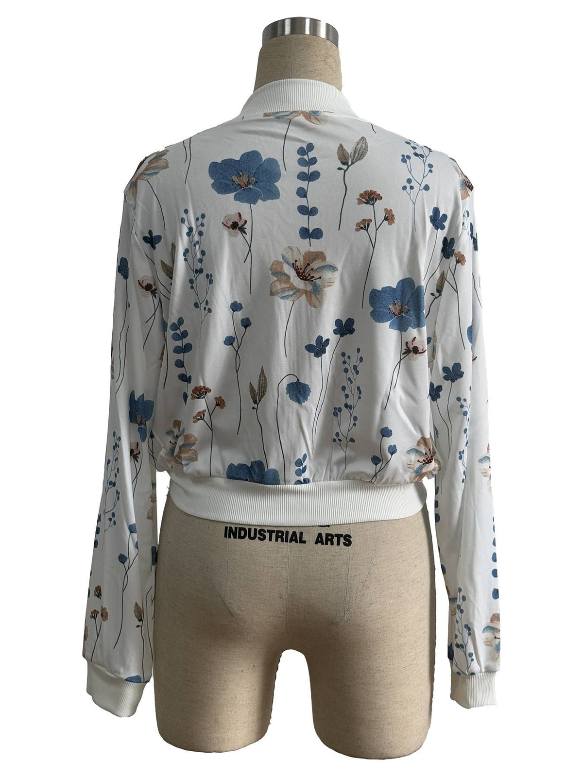 A white long-sleeve blouse jacket from the back with a blue and beige floral print, shown from the back on a beige dress form. The waistband of the blouse is elastic and sits above the black text 'INDUSTRIAL ARTS' printed on the mannequin's waist.