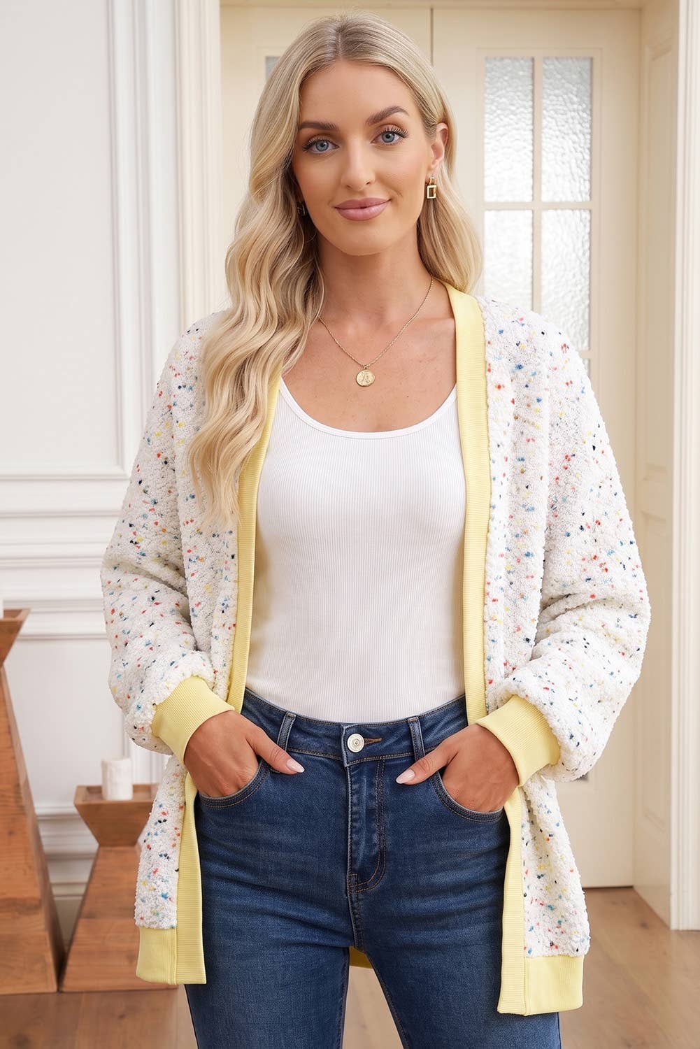 A woman modeling a cream-colored open-front cardigan with a delicate multicolor floral print, yellow ribbed trim, and a yellow collar, worn over a white tank top and blue jeans.