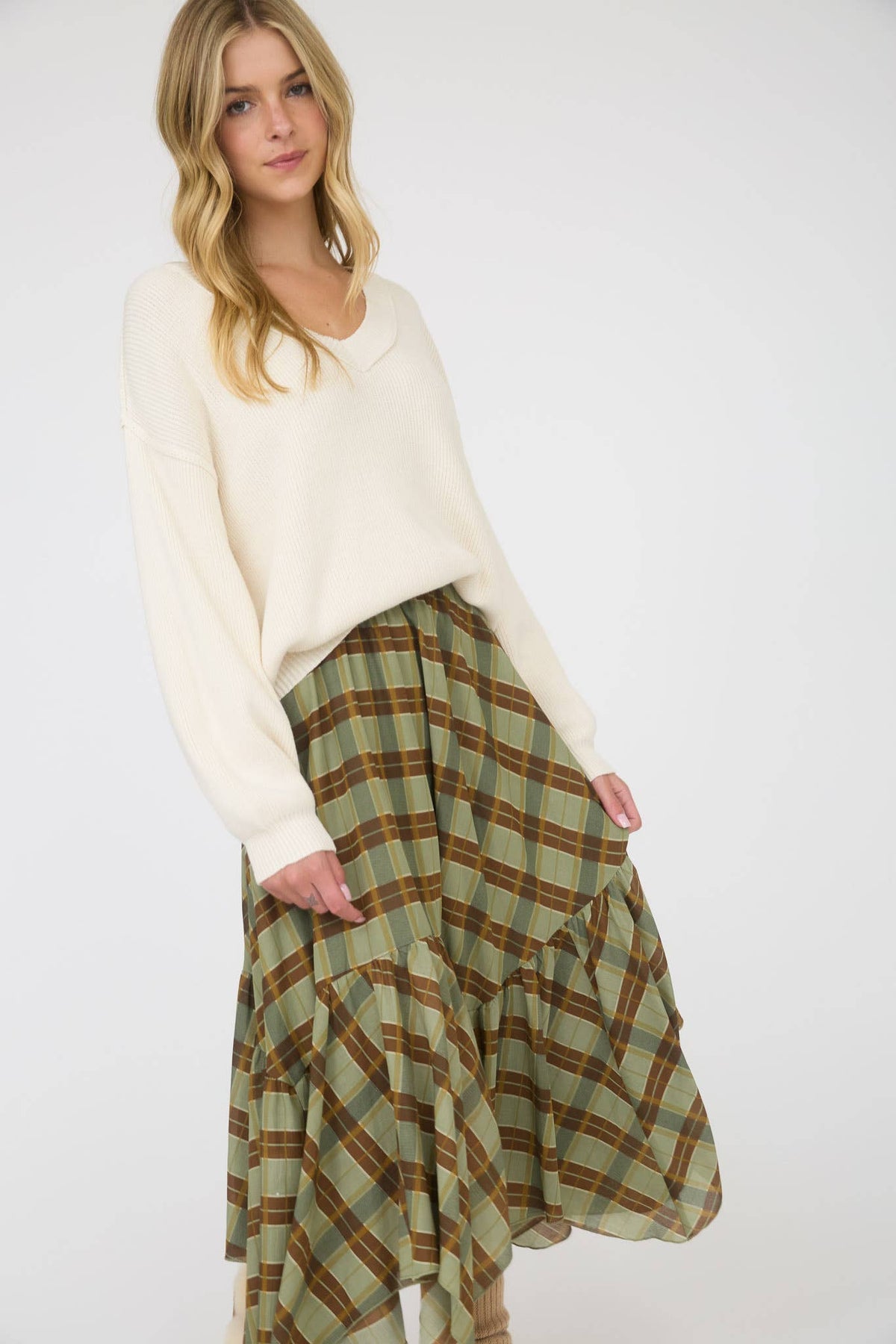 A woman modeling a cream-colored knit sweater and a green plaid skirt with brown and tan lines, set against a plain light gray studio background.