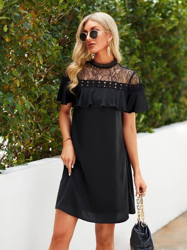 A woman wearing a black short-sleeve dress with a sheer lace yoke and ruffle trim, standing outdoors against a white wall and green hedge while holding a black handbag.