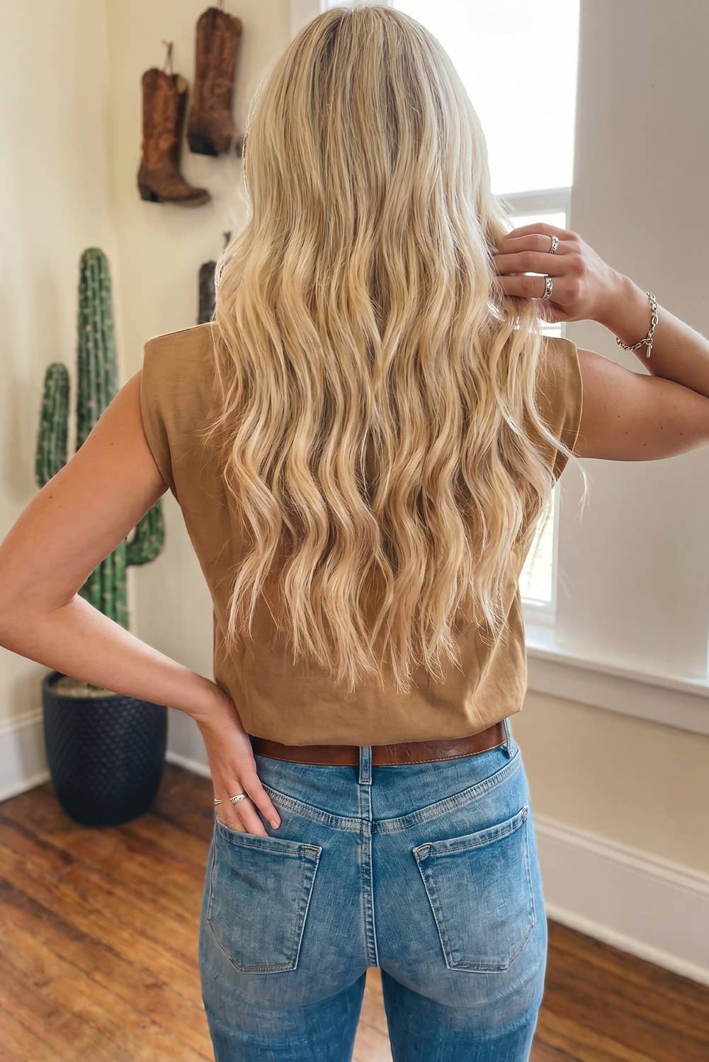 Person with long blonde hair wearing a brown top and blue jeans, standing in a room with a cactus plant and cowboy boots on the wall.