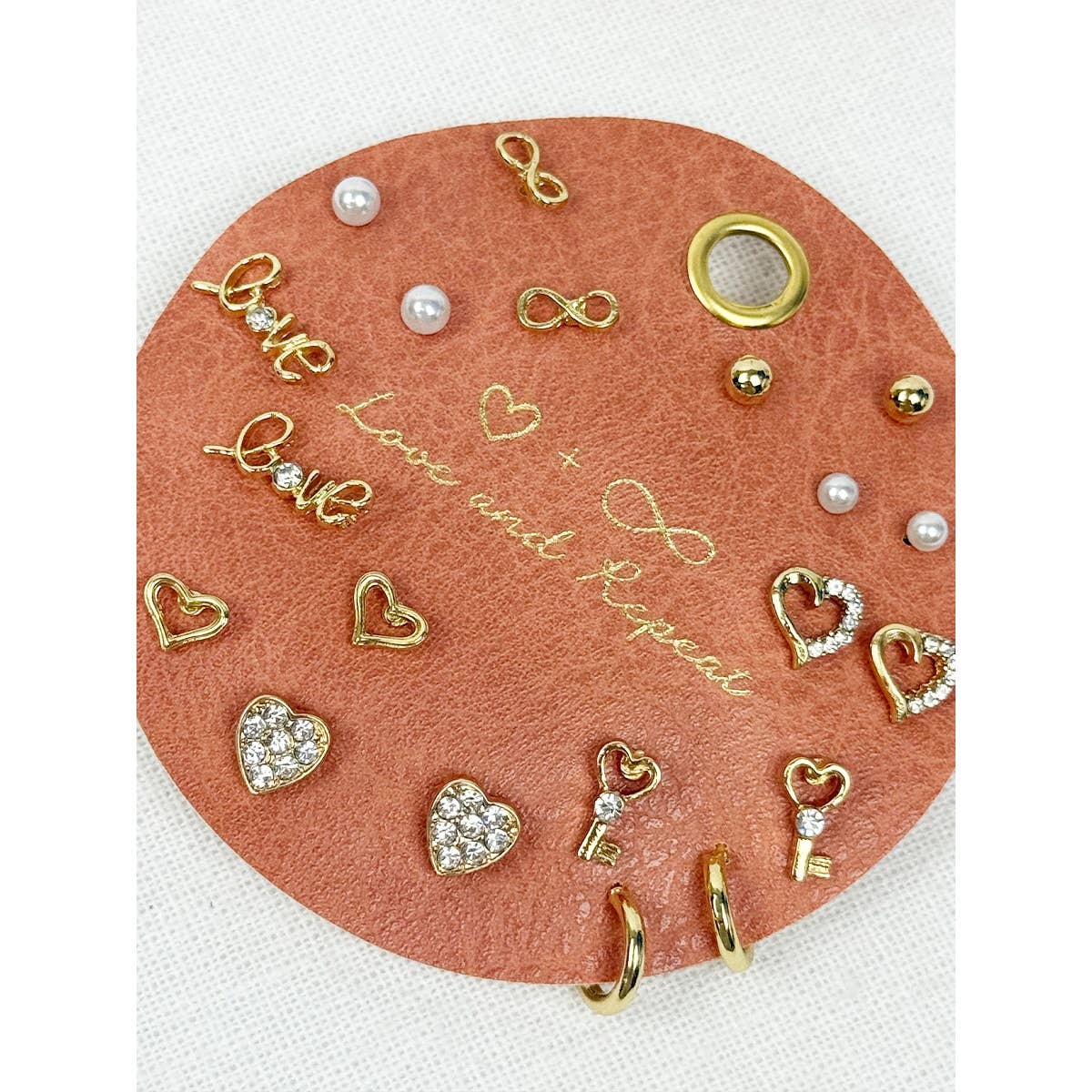 Collection of gold and pearl jewelry charms on a pink leather patch with text.