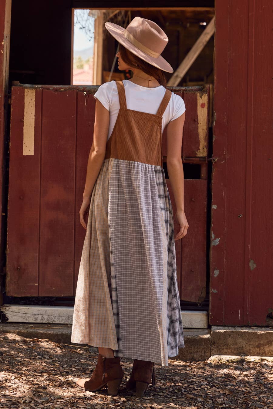 Woman wearing a brown overall dress over a white shirt and plaid skirt, standing in front of a red wooden door.