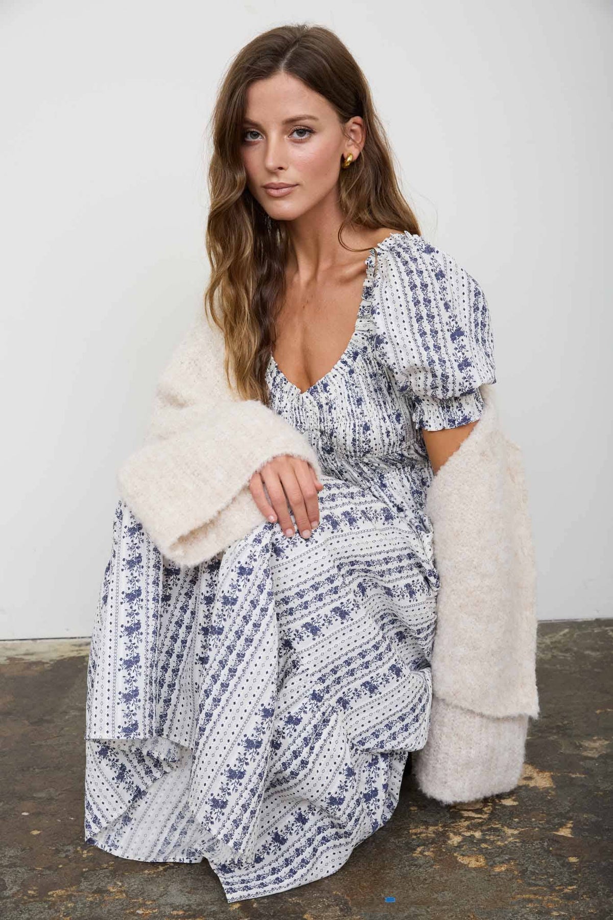A woman wearing a white and navy blue patterned maxi dress with puff sleeves and a fuzzy cream faux-fur cardigan, sitting against a plain light gray wall.