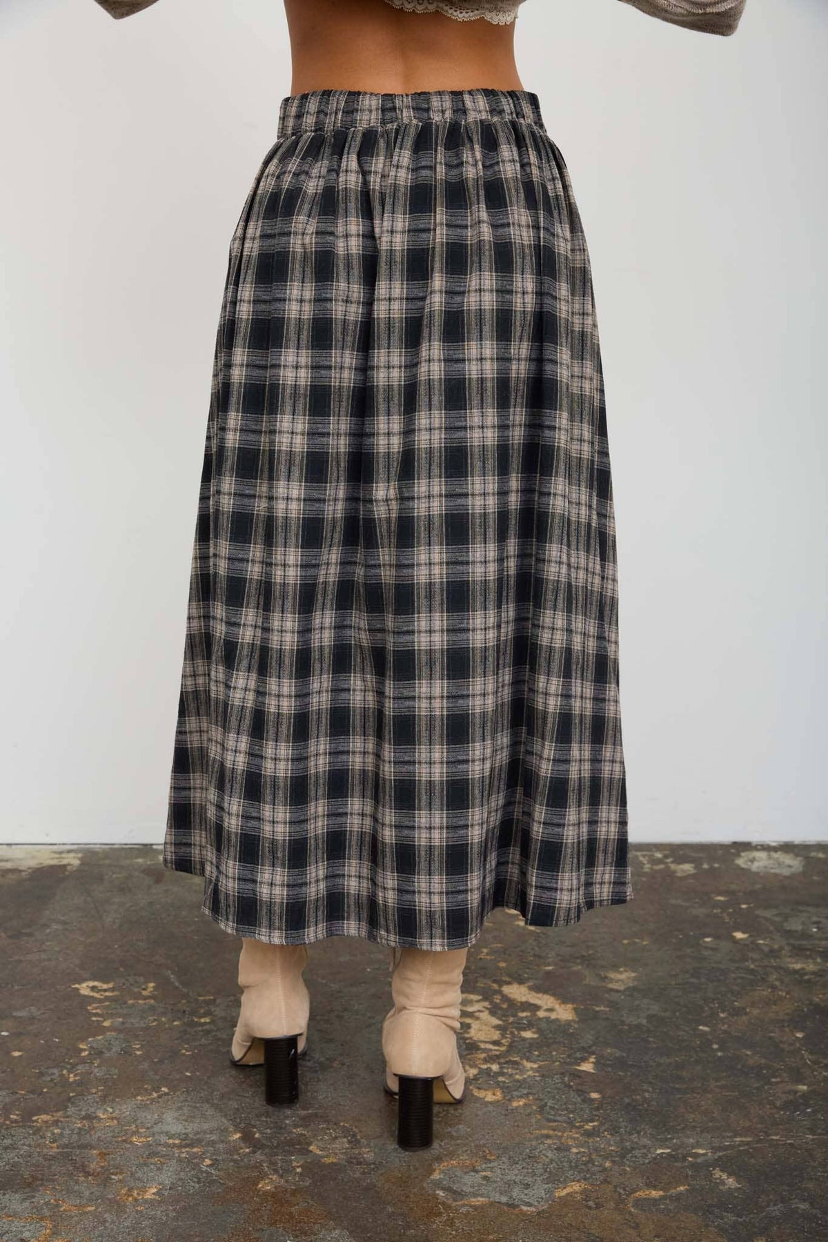 A person shown from the back wearing a long plaid midi skirt in dark navy, gray, and beige, paired with beige ankle boots and black heels, standing on a textured concrete floor against a plain white wall.