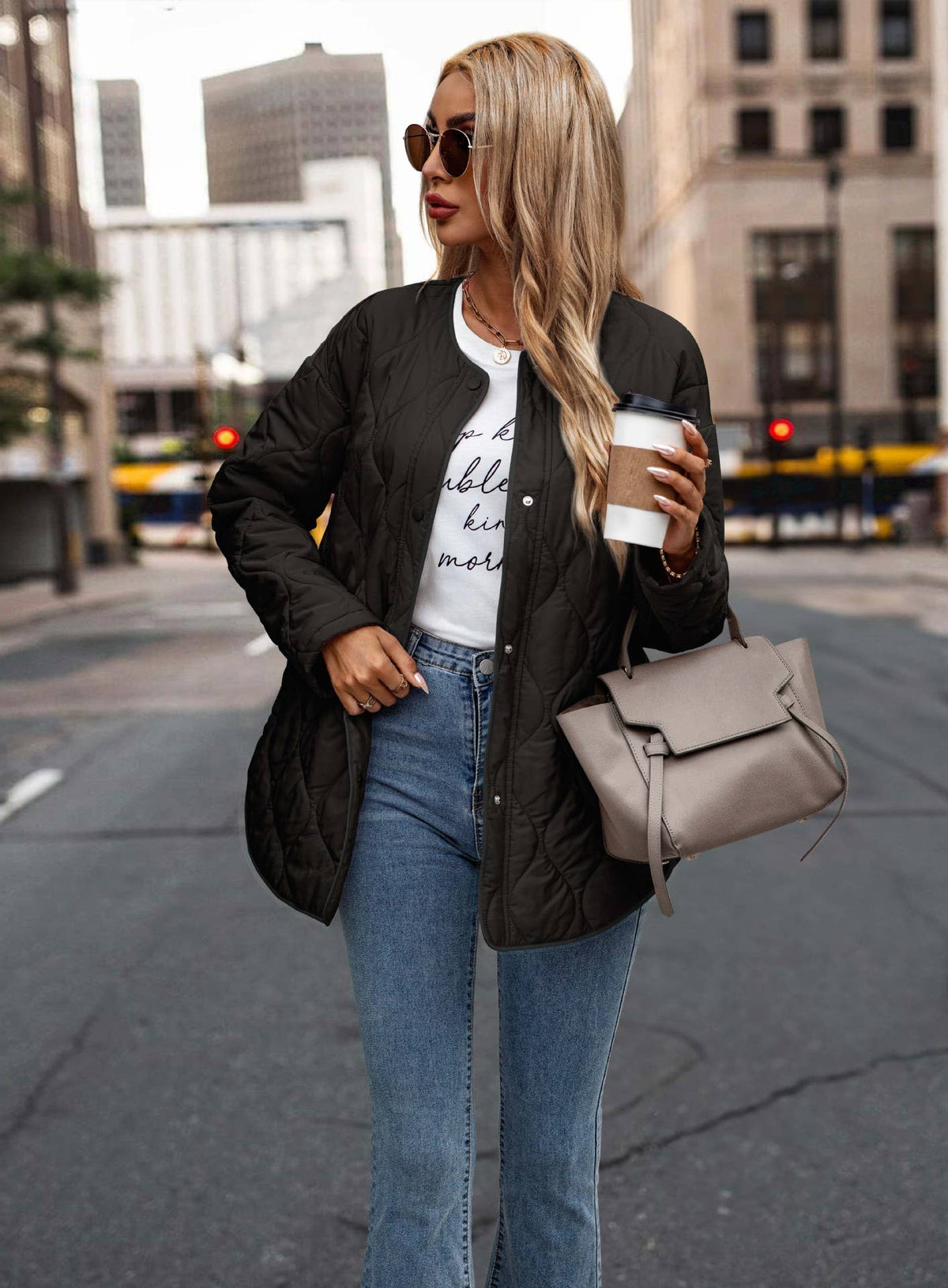 A woman standing on a city street wearing a black quilted bomber jacket, blue jeans, a white graphic t-shirt, sunglasses, and carrying a taupe-gray handbag and a coffee cup.