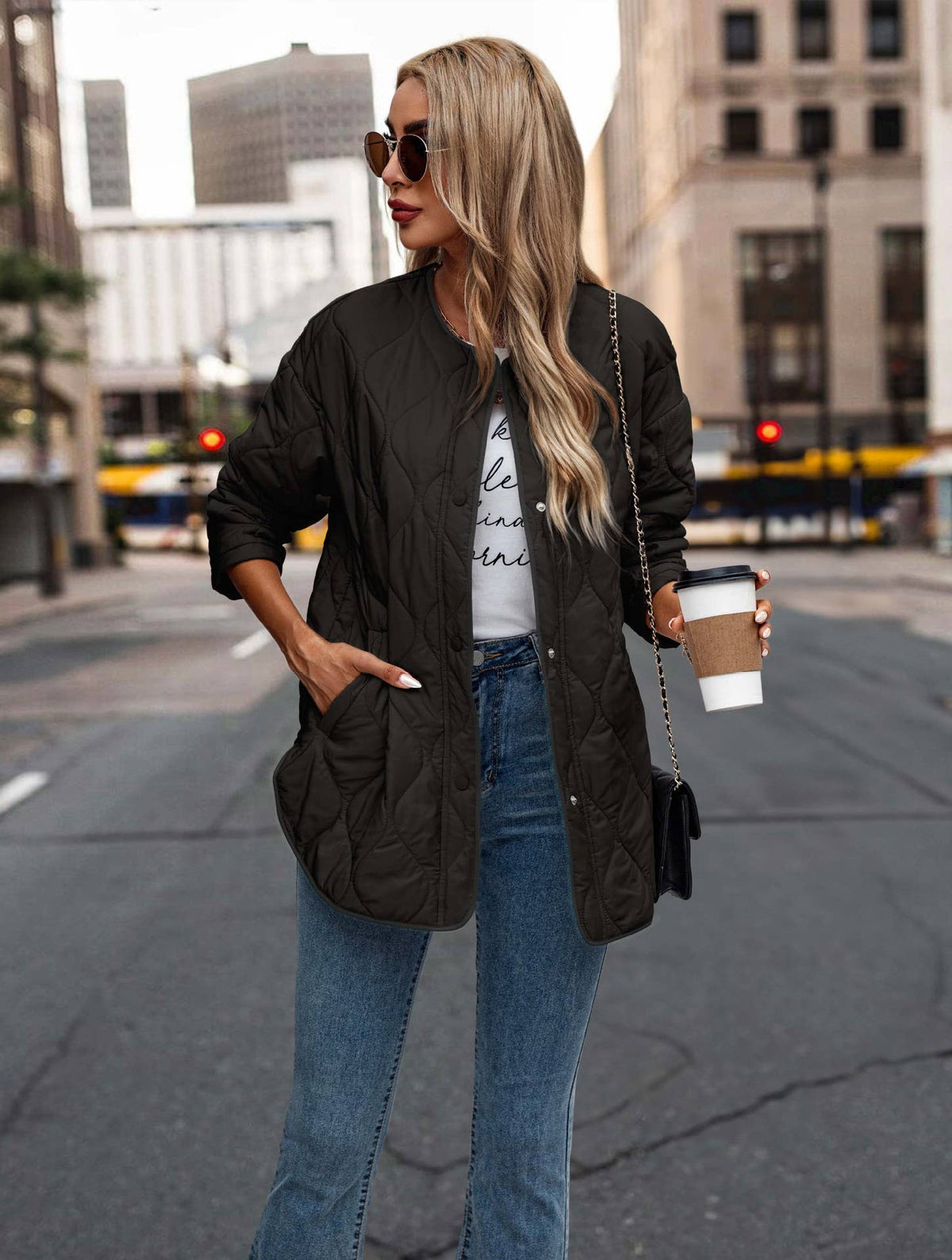 A woman wearing a black quilted zip-up jacket, blue jeans, and sunglasses stands on a city street holding a coffee cup.