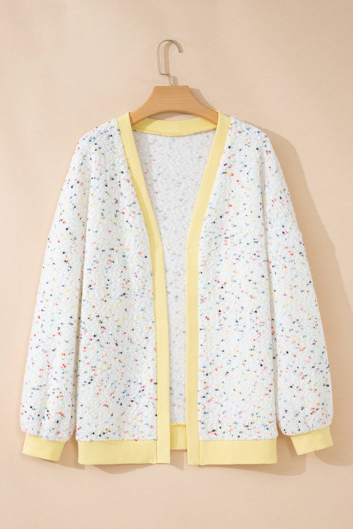 A white open-front cardigan with a multicolor speckled pattern and light yellow trim, hanging on a wooden hanger against a solid beige background.
