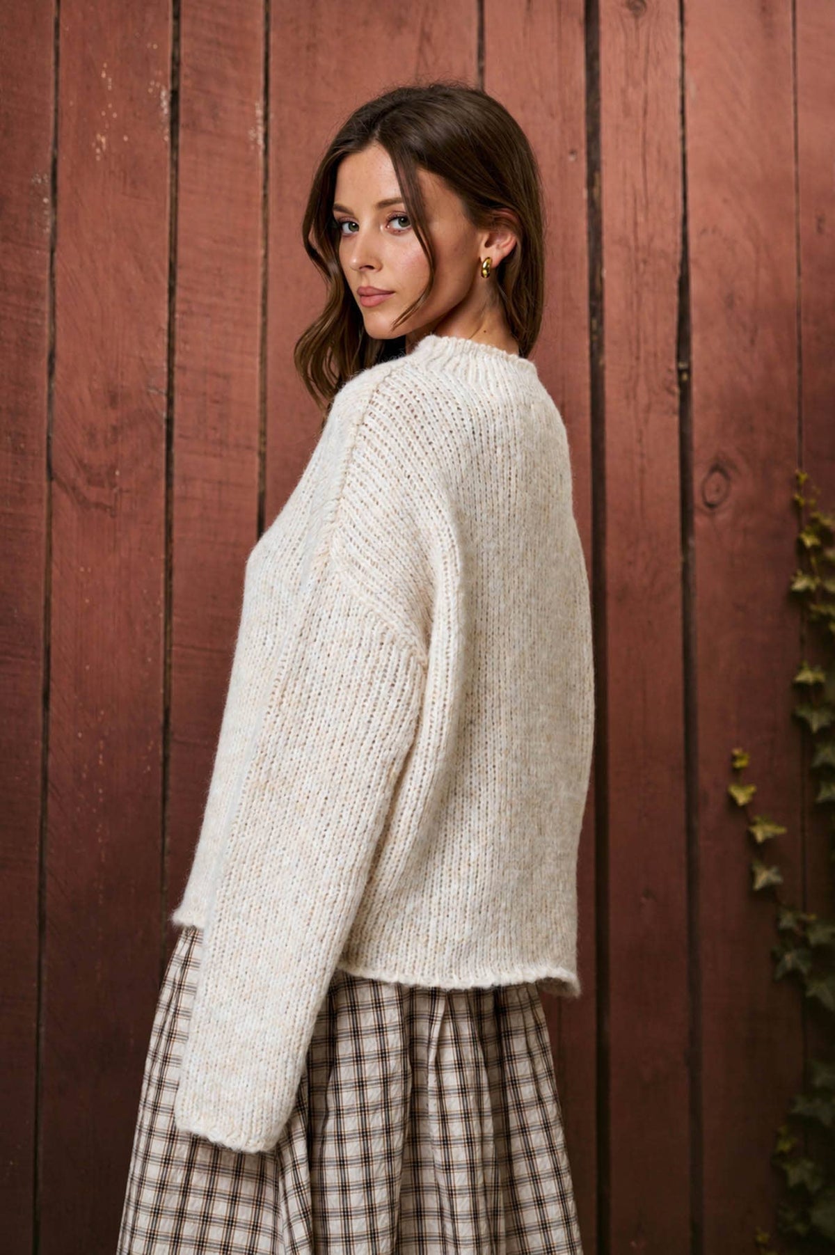 Woman wearing a white sweater and plaid skirt standing against a wooden fence.