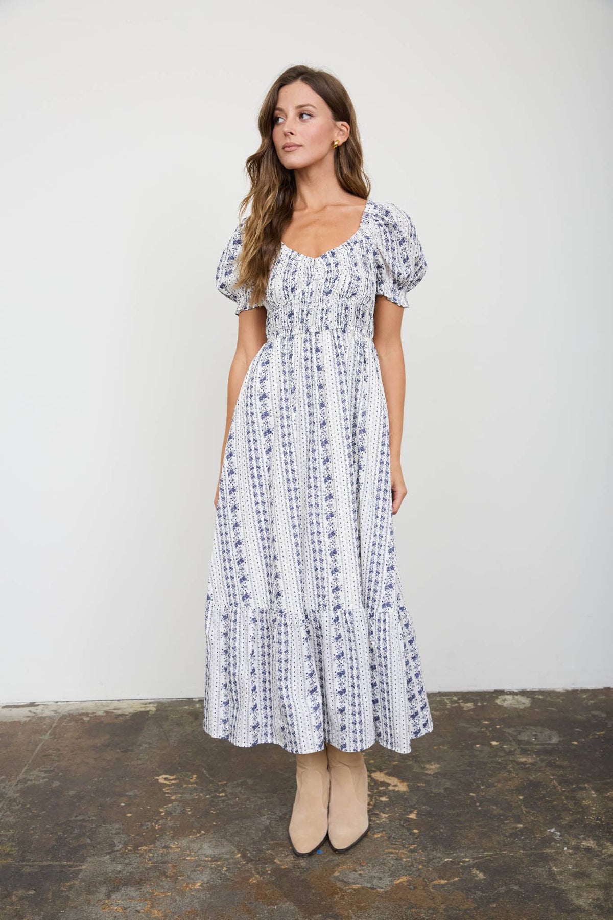A woman modeling a white midi dress with a blue vertical floral pattern, puff sleeves, and a smocked bodice, standing against a plain white wall.