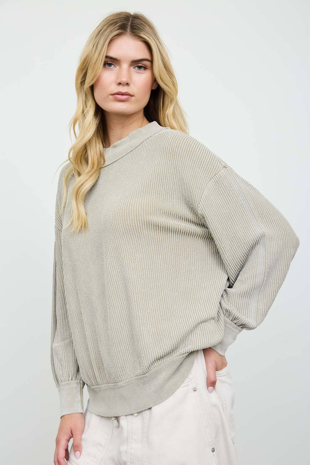 A blonde model wearing a light gray ribbed knit long-sleeve top and white pants, standing against a light gray studio background.