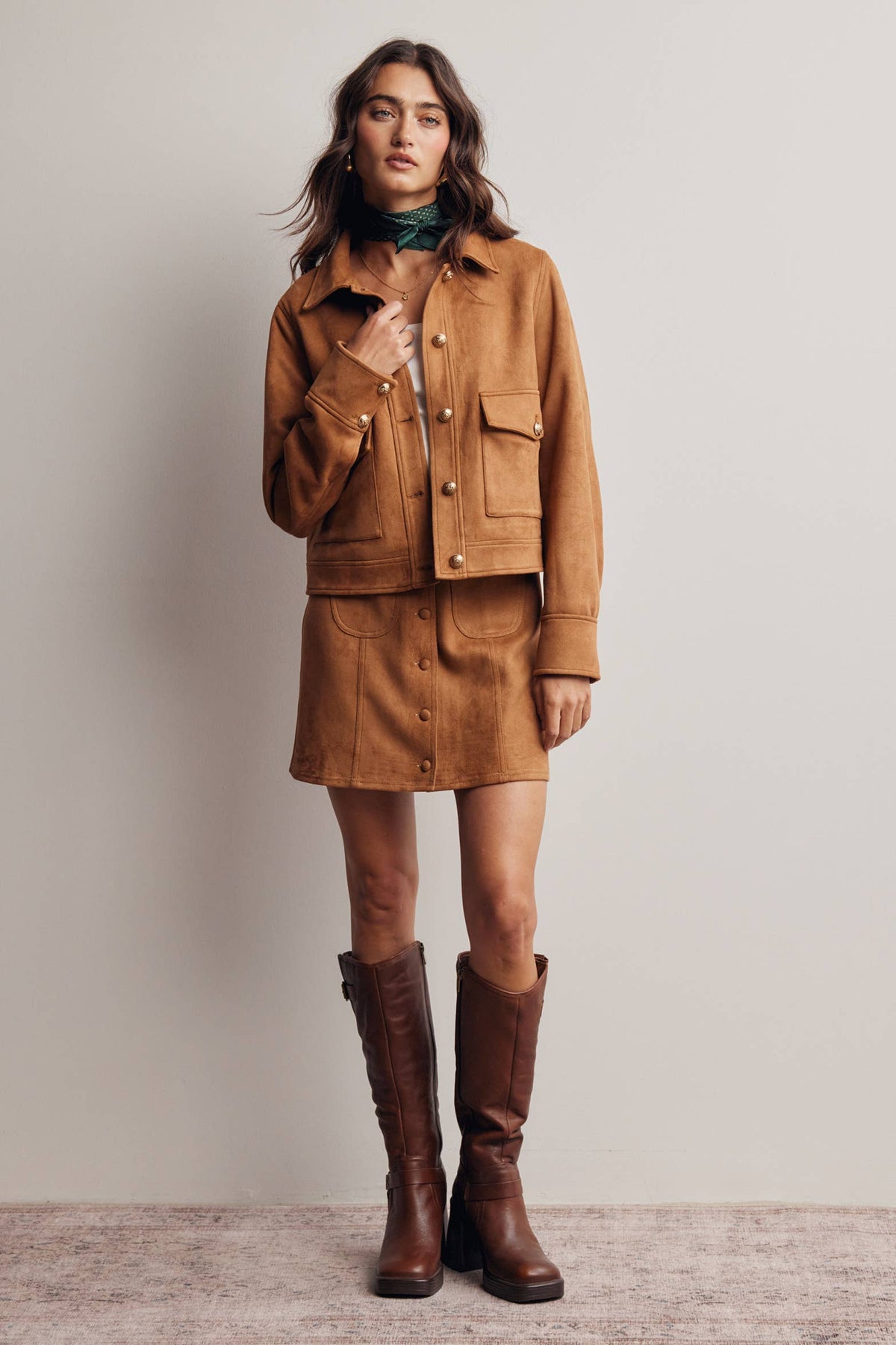 Woman wearing a brown suede jacket and skirt with knee-high boots on a plain background