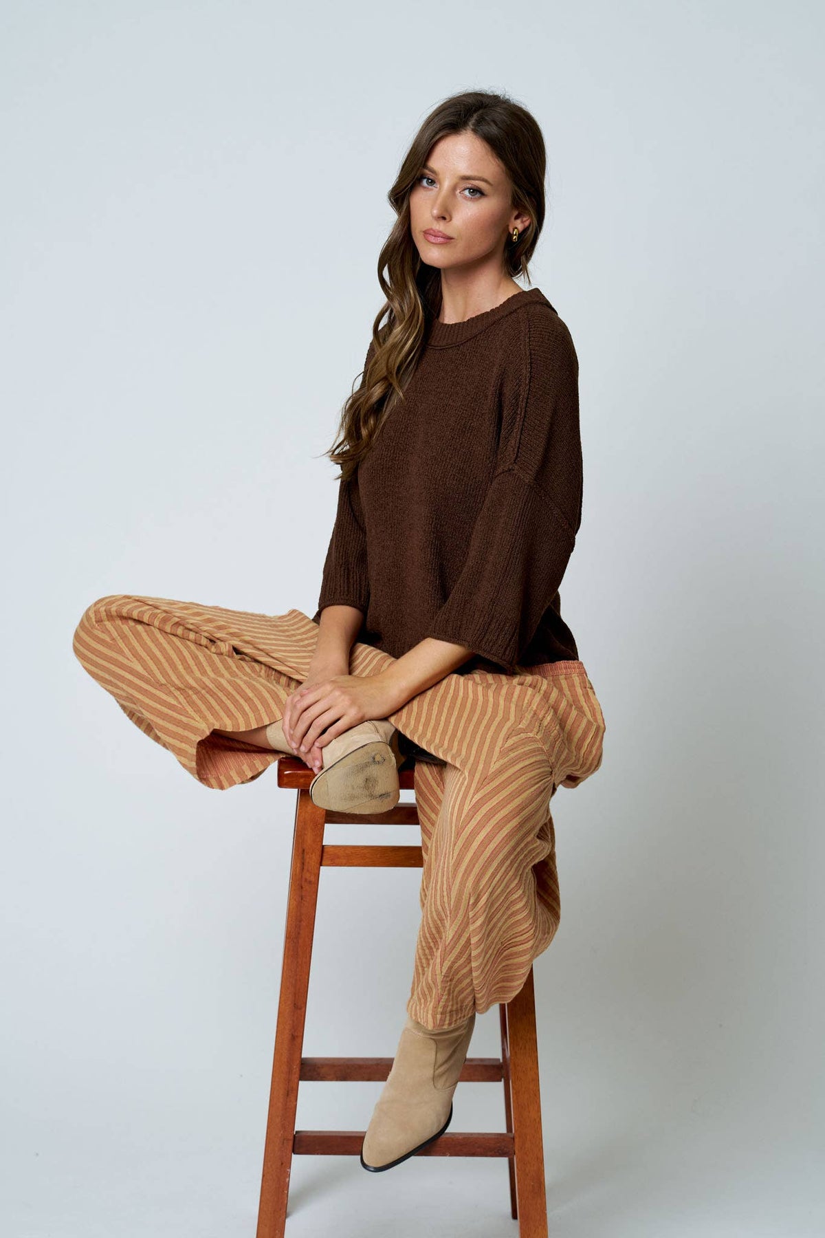 Woman sitting on a wooden stool wearing a brown sweater and beige pants against a white background