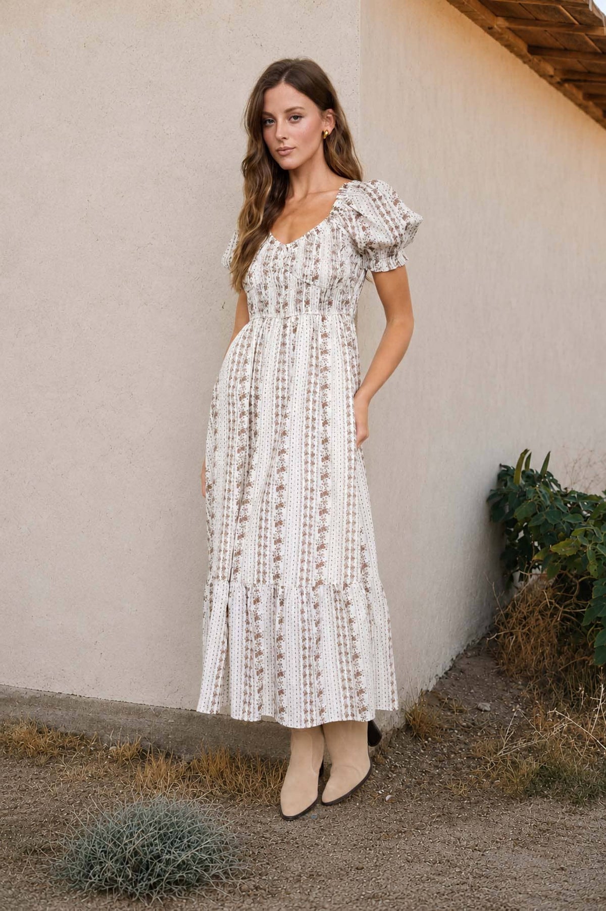 A woman modeling a white maxi dress with a delicate vertical floral pattern, featuring ruffled short sleeves and a smocked bodice, standing against a beige stucco wall.