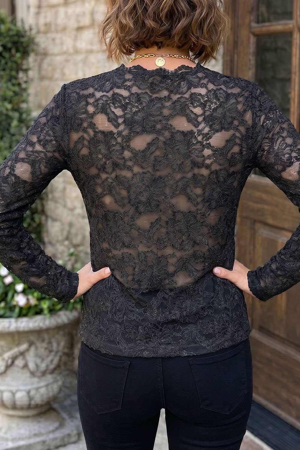 A woman viewed from the back wearing a sheer black lace long-sleeve top and black pants, standing outdoors in front of a stone wall and wooden door.