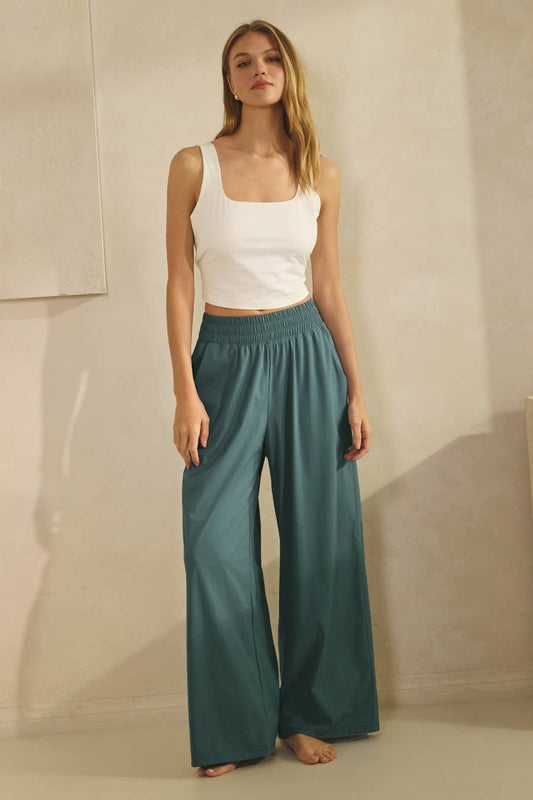 Woman wearing a white tank top and teal pants against a neutral background
