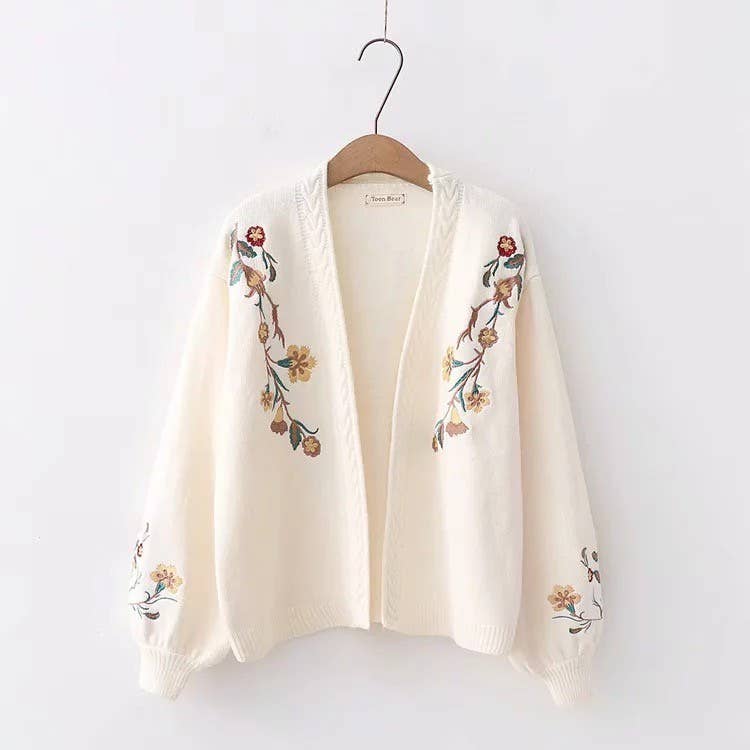 White cardigan with floral embroidery on a wooden hanger against a white background