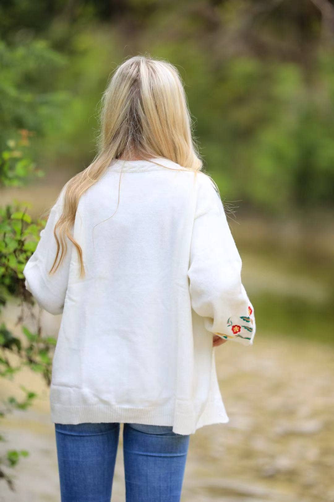 Person wearing a white sweater with a floral design on the sleeve, standing outdoors.