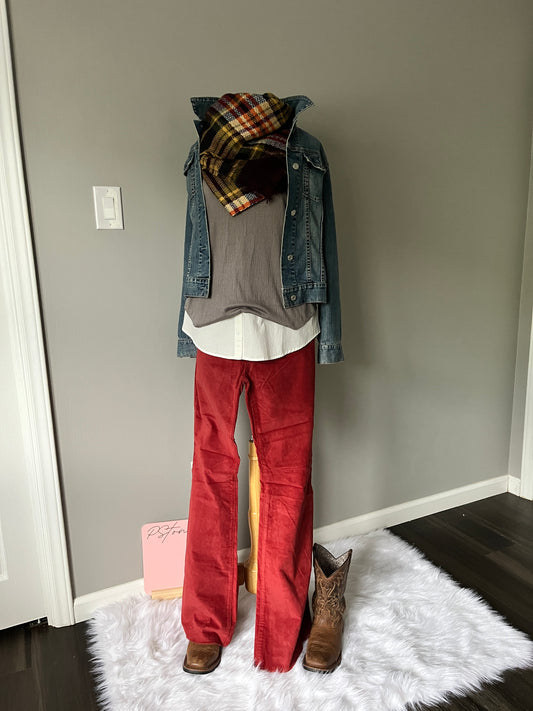 Bare wall, hardwood floors and white shag rug. Mannequin styled with a plaid scarf, denim jacket in medium wash, grey top, white button shirt peeking out, and rust corduroy flare pants with brown cowboy boots.