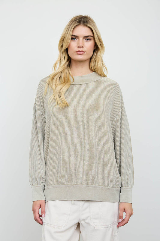 A blonde woman wearing a light beige ribbed knit long-sleeve sweater and white pants, standing against a light gray studio background.