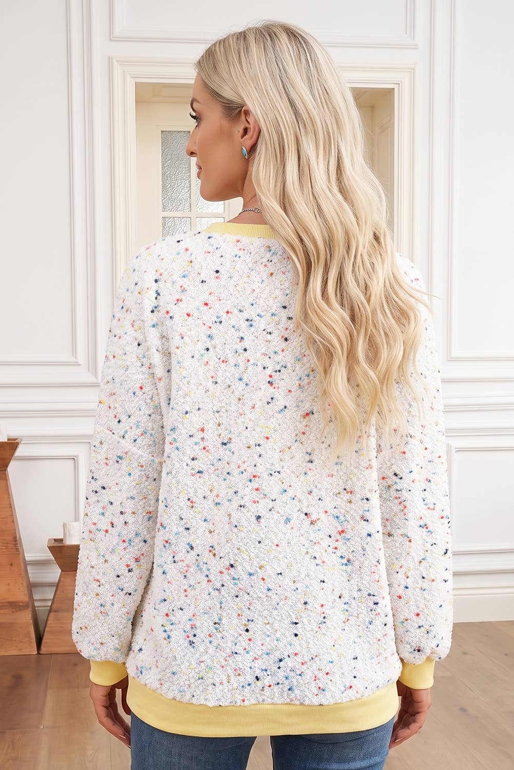 A woman with long blonde hair is shown from the back wearing a white long-sleeve sweatshirt covered in a multicolor speckled pattern, with yellow ribbed trim at the collar, cuffs, and hem. She is standing in a bright room with white paneled walls and wood flooring.