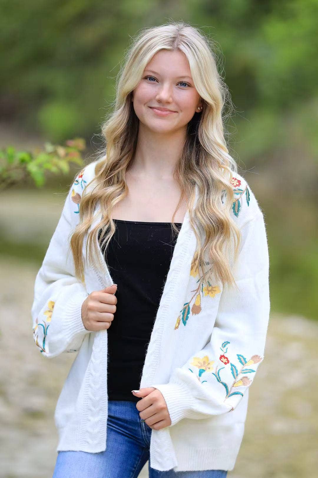 Woman wearing a white embroidered cardigan over a black top and blue jeans, standing outdoors with greenery in the background.