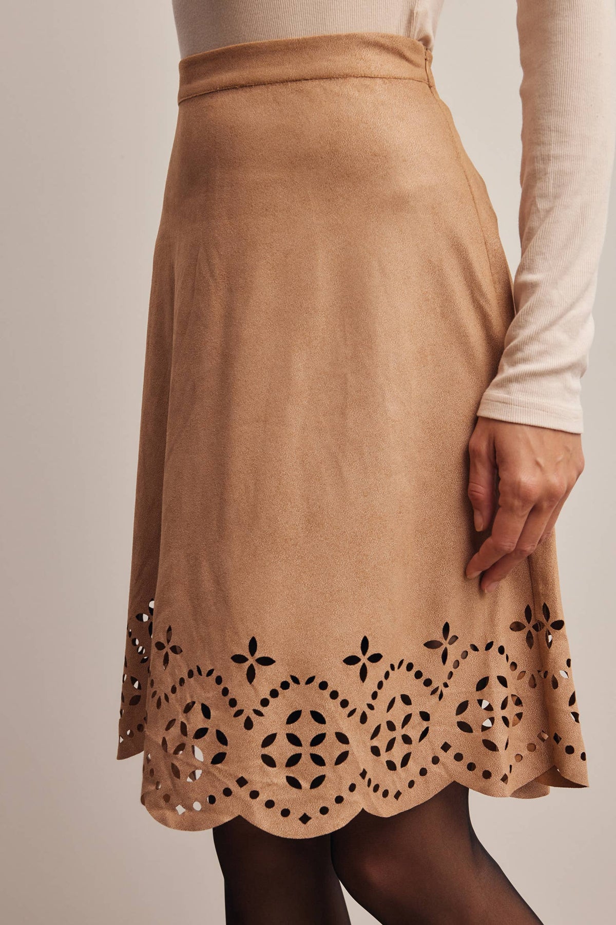 A tan A-line skirt with a scalloped eyelet lace hem, worn by a model in a cream long-sleeve top and black tights against a warm beige background.