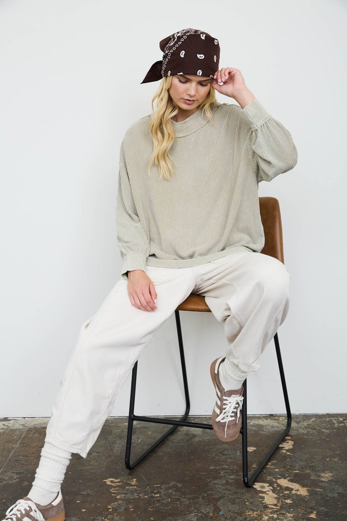 A woman sits on a brown chair wearing a beige knit sweater, white sweatpants, a brown bandana, and brown sneakers against a plain white wall.