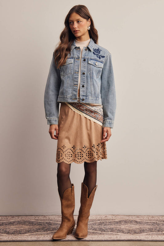 A woman stands against a plain beige wall wearing a light-wash denim jacket with blue floral embroidery on the chest, a tan patterned wrap skirt with a scalloped hem, dark tights, and brown cowboy boots.