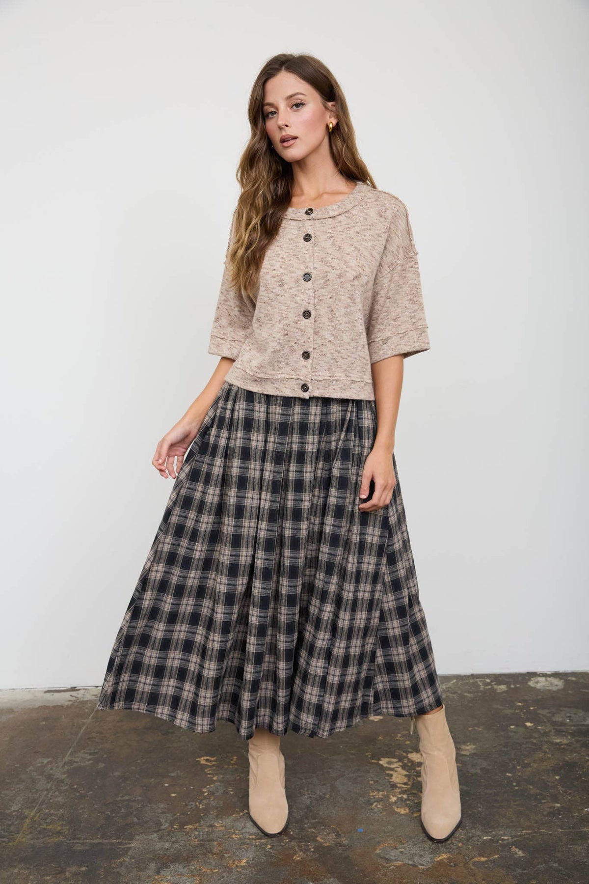 A woman modeling a two-piece outfit consisting of a beige short-sleeve cropped cardigan with buttons and a dark gray and black plaid midi skirt, paired with beige ankle boots, set against a plain light gray wall and concrete floor.