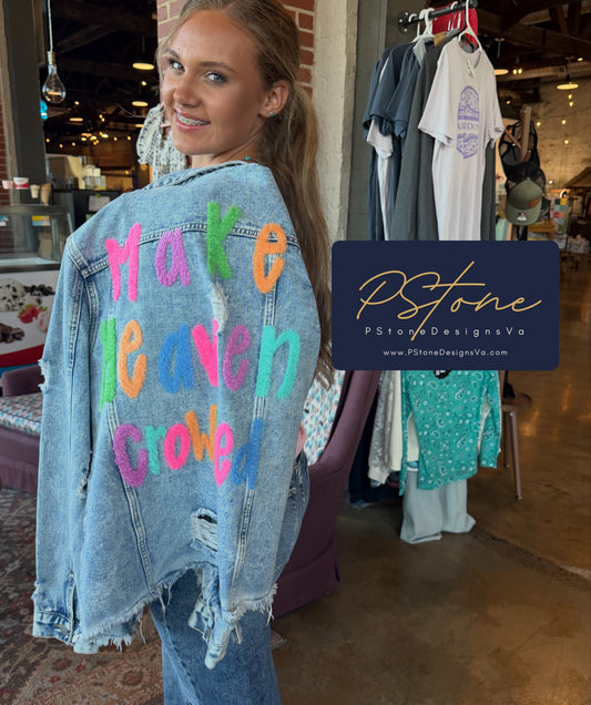 A woman wearing a light-wash distressed denim jacket with colorful embroidered text reading 'Make Heaven Crowded' in a boutique setting. A dark blue brand overlay for 'PStone Designs by PStone' is positioned on the right side of the image.