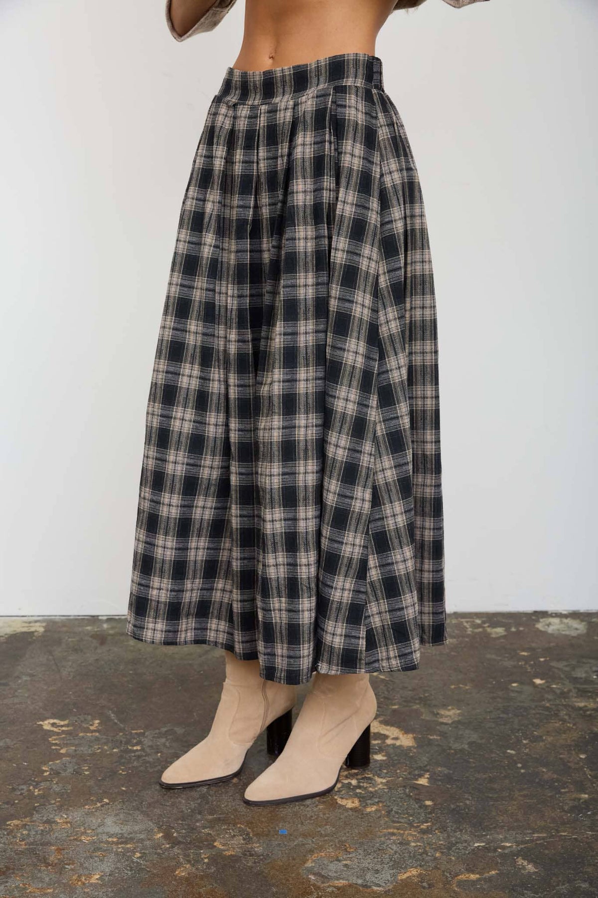 A person wearing a high-waisted, wide-leg plaid midi skirt in black, gray, and beige, paired with beige ankle boots, standing on a textured concrete floor against a plain white wall.