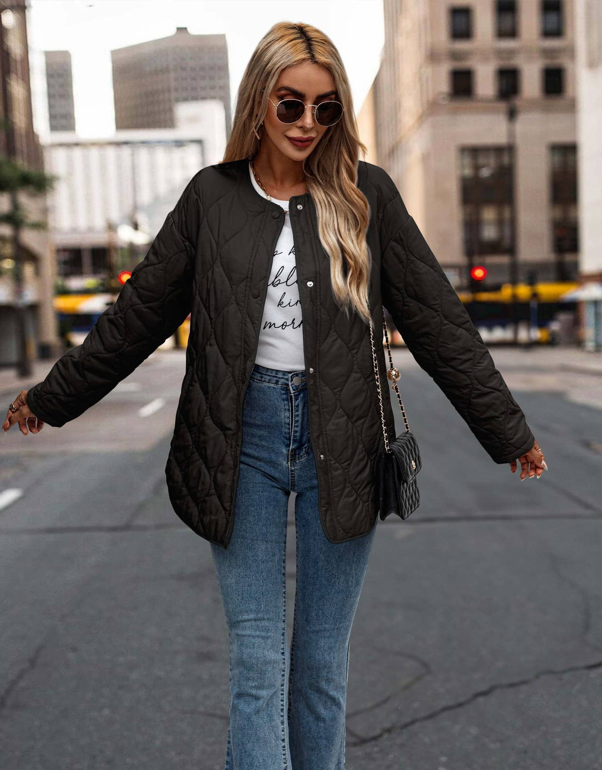 A woman wearing a black quilted bomber jacket over a white graphic t-shirt and blue jeans, walking on a city street.
