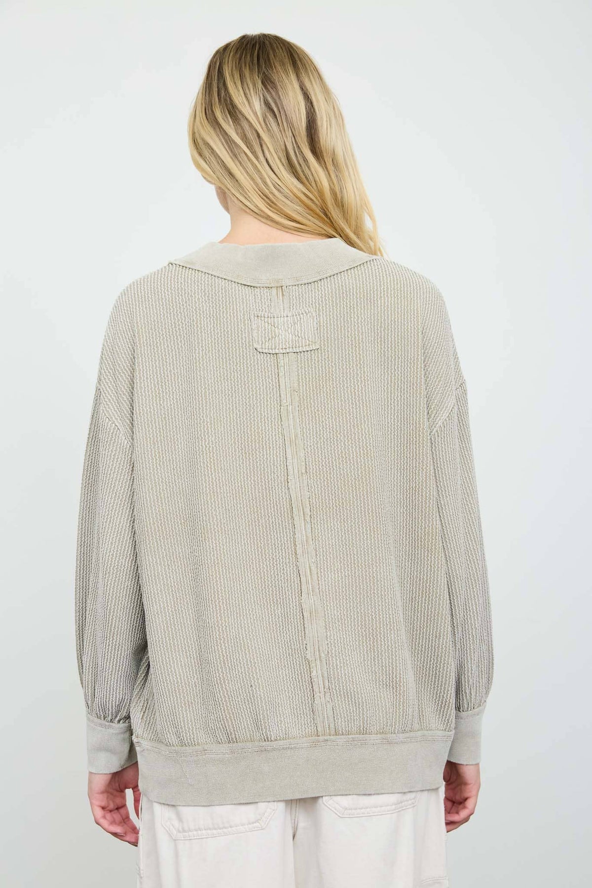 A woman viewed from the back wearing a light beige ribbed knit sweater with a unique patch detail on the upper back, paired with white pants against a plain white background.