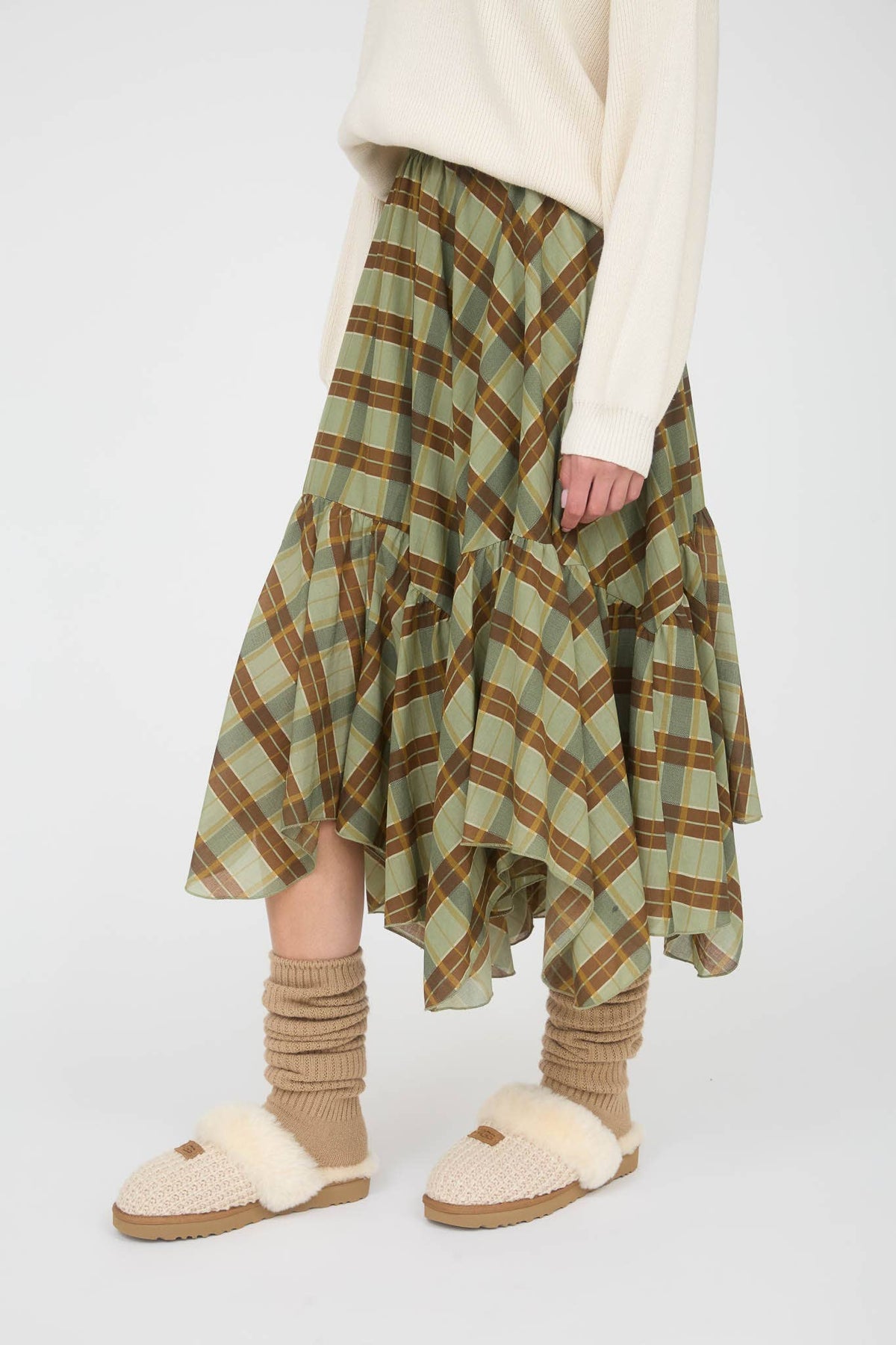 A person wearing a green and brown plaid tiered skirt with an asymmetrical hem, styled with a cream sweater, beige socks, and knit slippers against a light gray studio background.