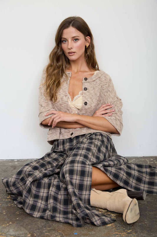 Woman wearing a beige sweater and plaid skirt sitting against a white wall.