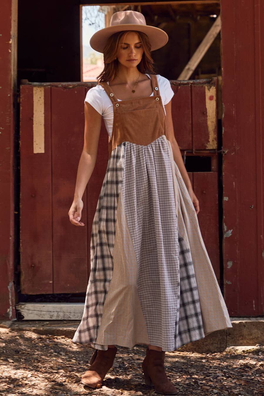 Woman wearing a brown overall dress over a white shirt and plaid skirt, standing in front of a red wooden door.