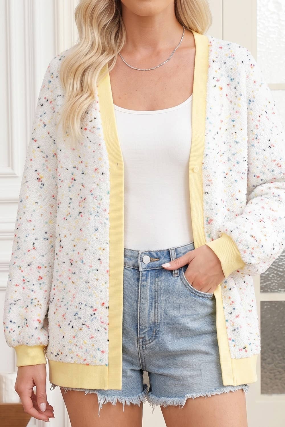 A woman wearing a cream cardigan with a multicolor speckle pattern and yellow trim, styled over a white tank top and denim shorts.