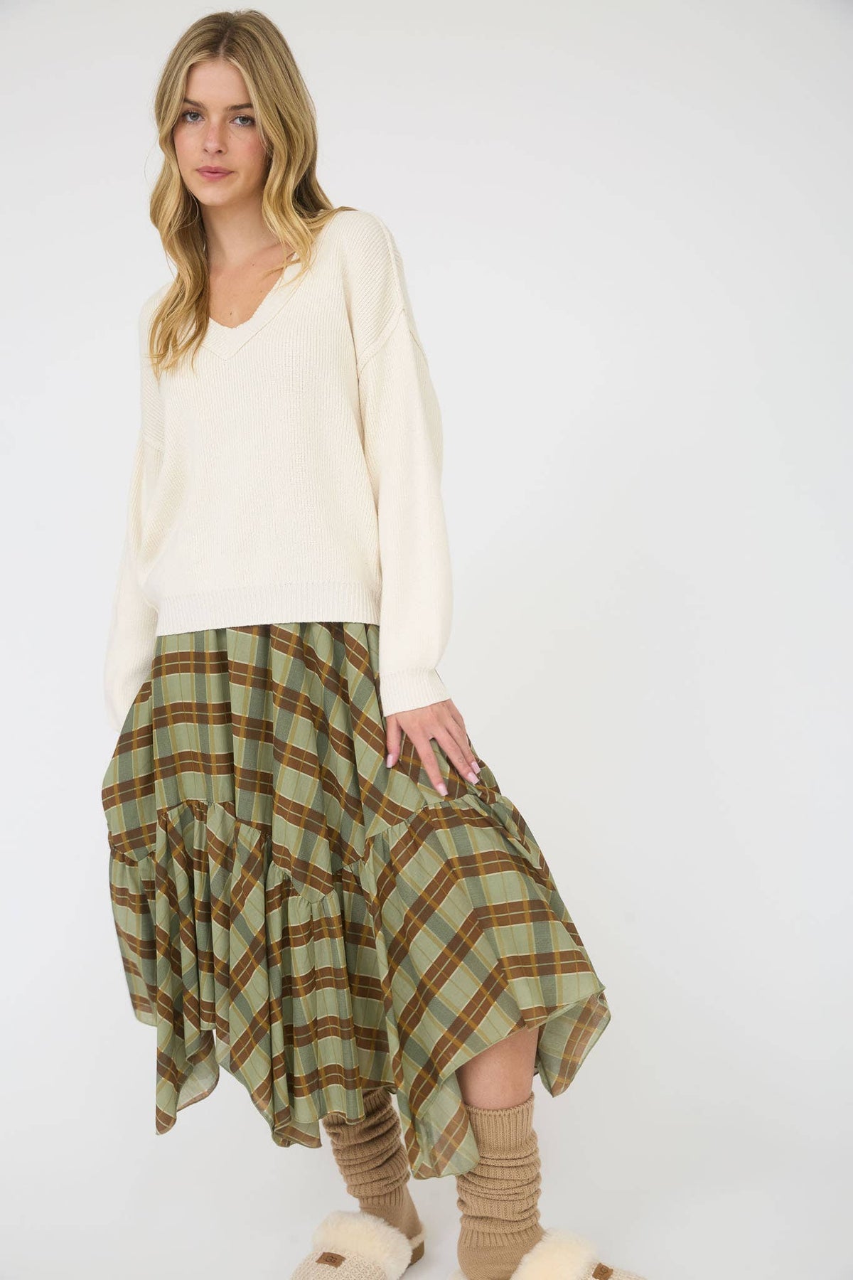 A woman modeling a cream-colored knit sweater and a green and brown plaid tiered skirt, paired with beige socks and fluffy slippers, against a plain white studio background.