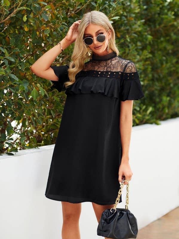 A woman wearing a black short-sleeve dress with a sheer lace yoke and ruffled trim, standing outdoors against a white wall and green foliage. She is holding a black handbag with a gold chain strap.