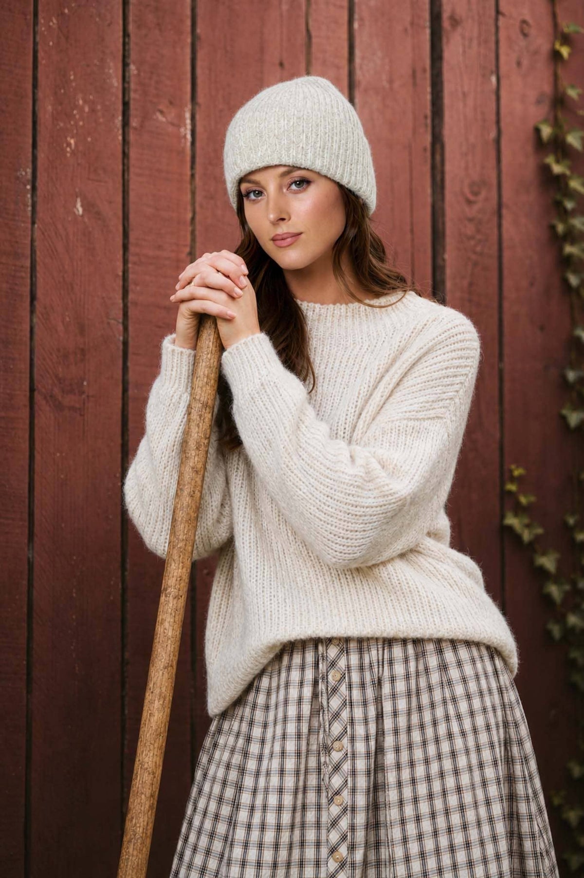 Woman wearing a white knit sweater and beanie holding a broom against a wooden wall.