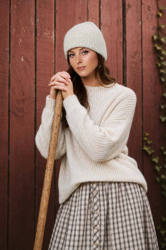 Woman wearing a white knit sweater and beanie holding a broom against a wooden wall.
