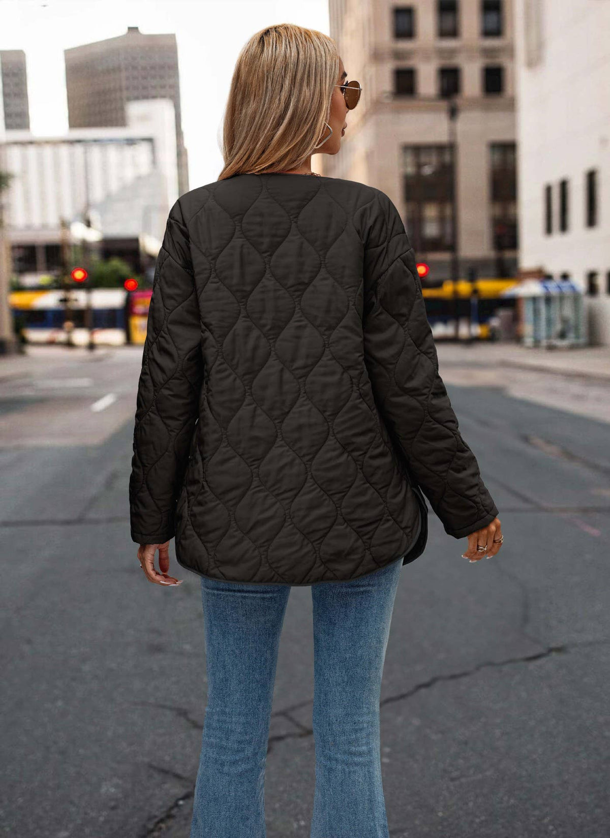 A woman viewed from behind wearing a black diamond-quilted jacket and blue jeans, standing on a city street with buildings in the background.