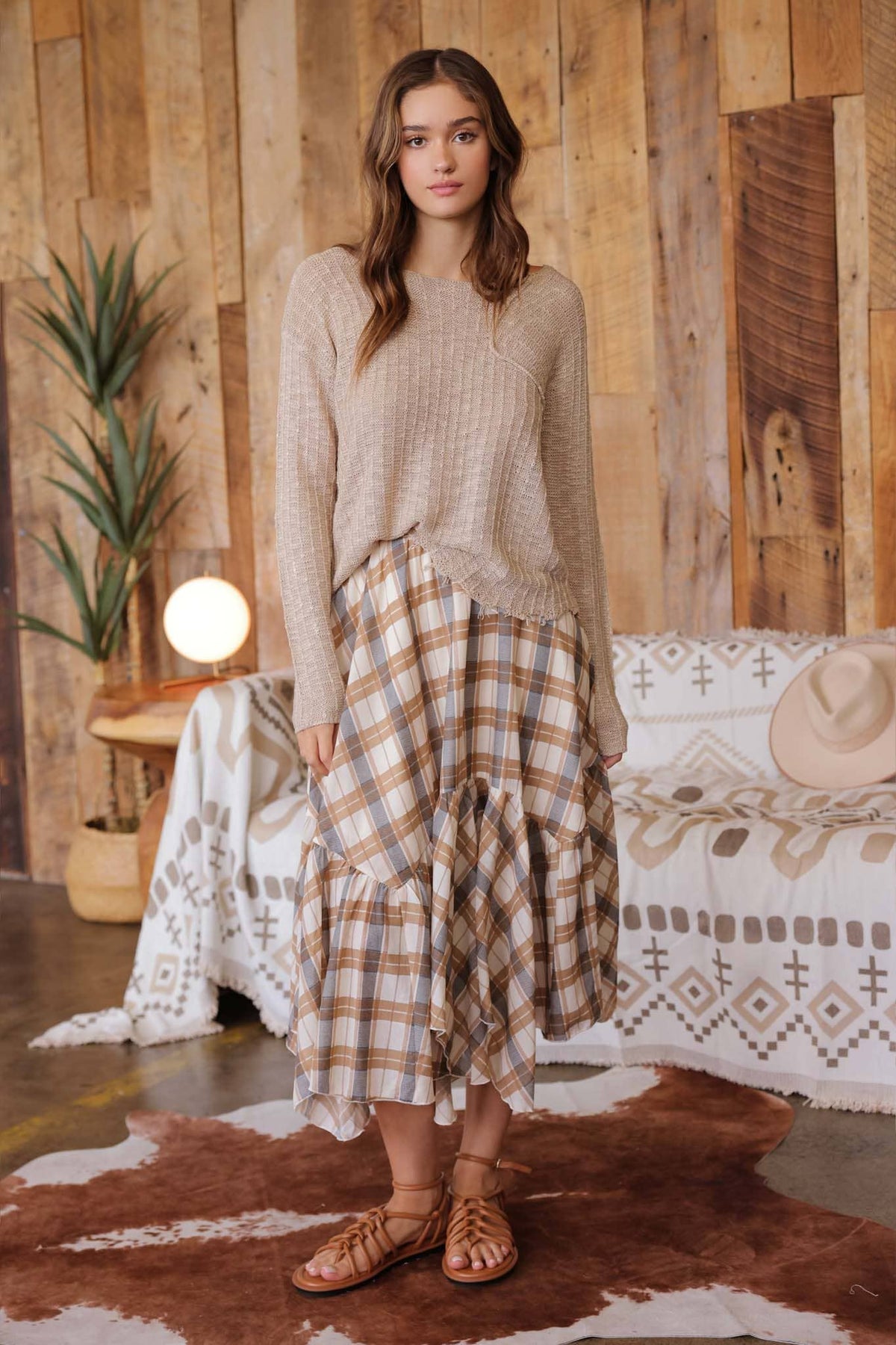 A woman stands in a rustic room wearing a beige knit long-sleeve top and a brown, beige, and white plaid tiered midi skirt, paired with brown leather sandals.