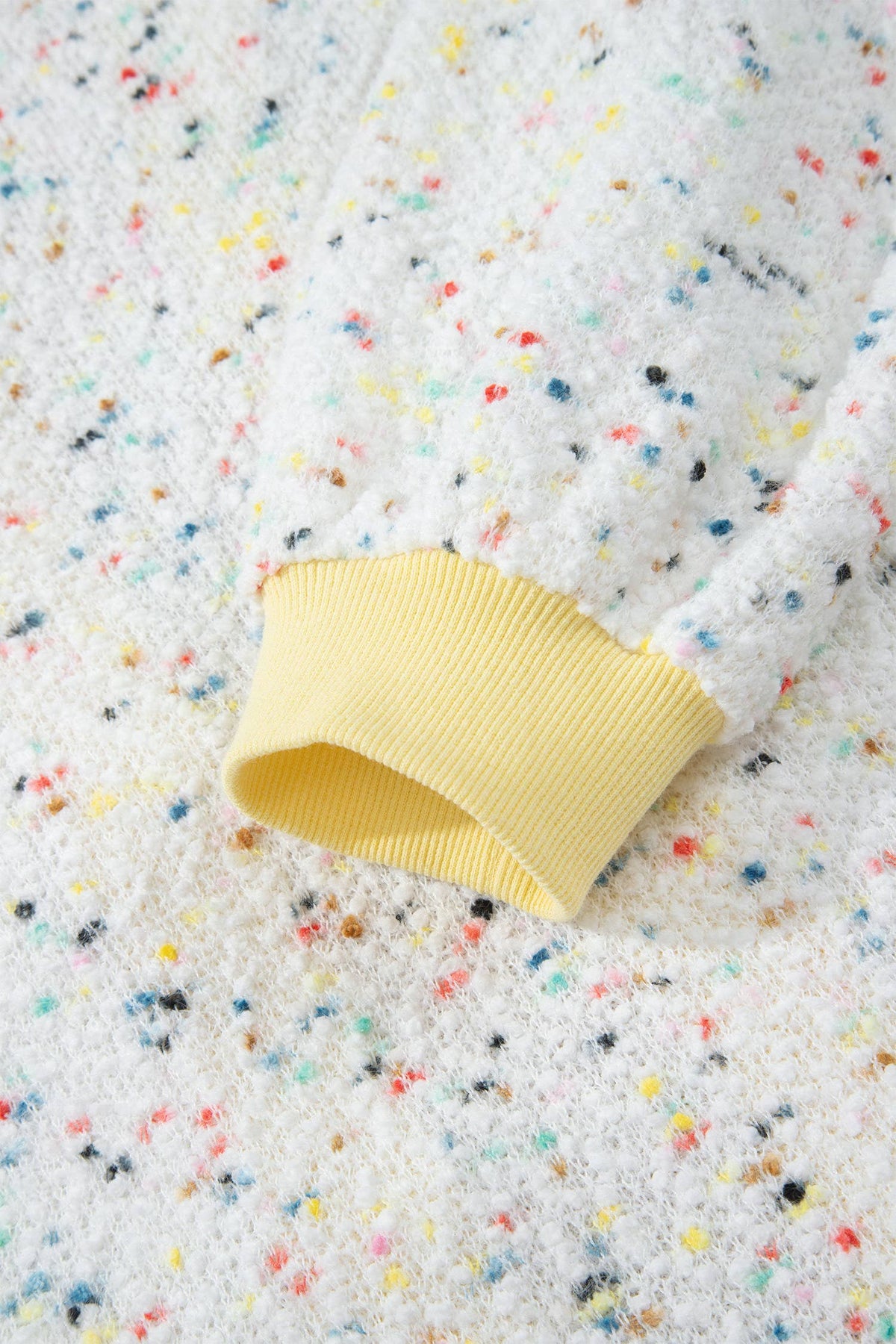 Close-up of a white fuzzy knit cardigan with multicolored confetti speckles and a soft yellow ribbed cuff.