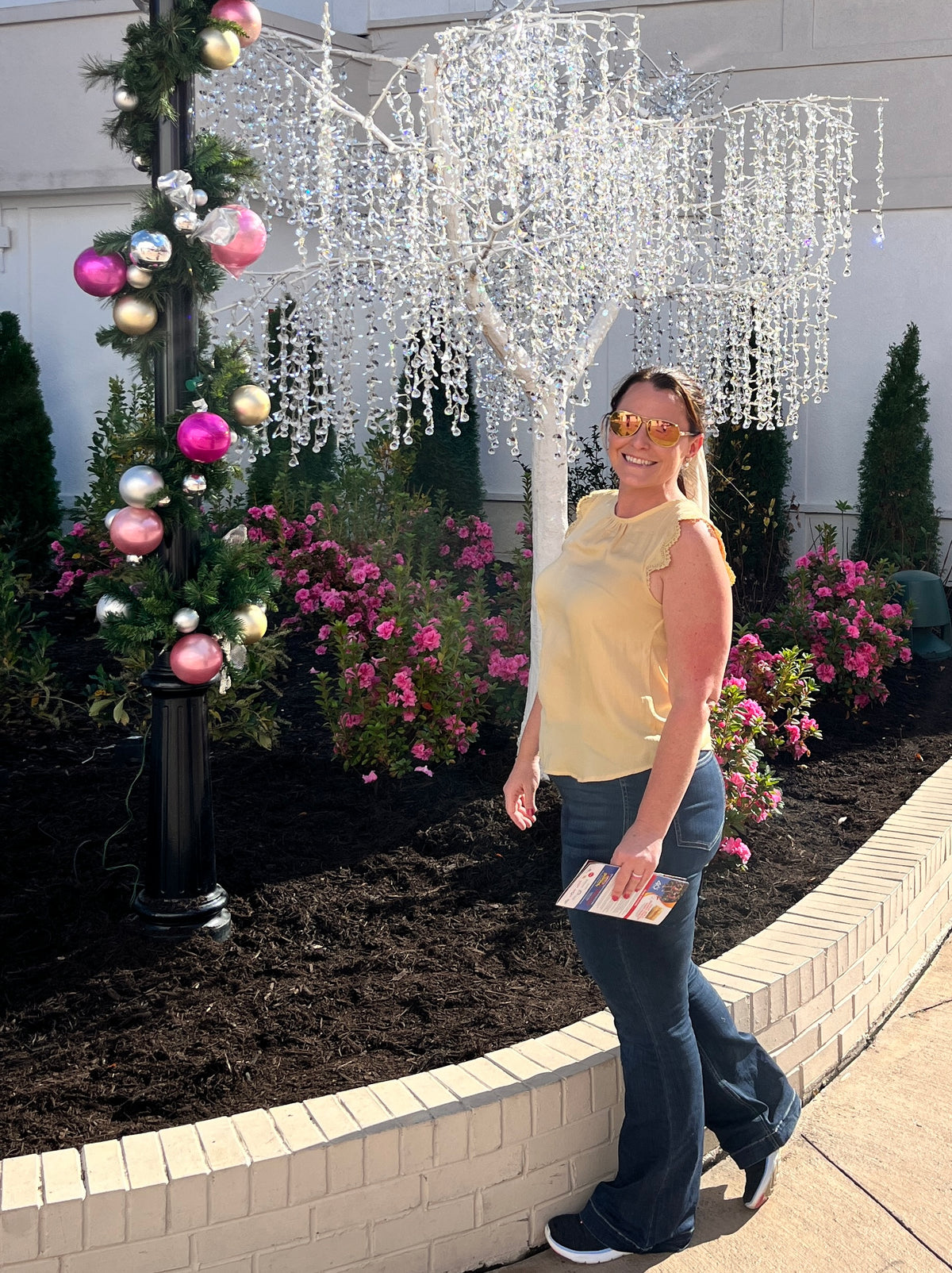 Image of PStone, owner of PStoneDesignsVa LLC, outside at Dollywood in front of an icicle tree, pink floers, and pink and silver holiday decorated garland. PStone is wearing dark wash pull on jeans, yellow sleeveless top, black sneakers, and sunglasses.