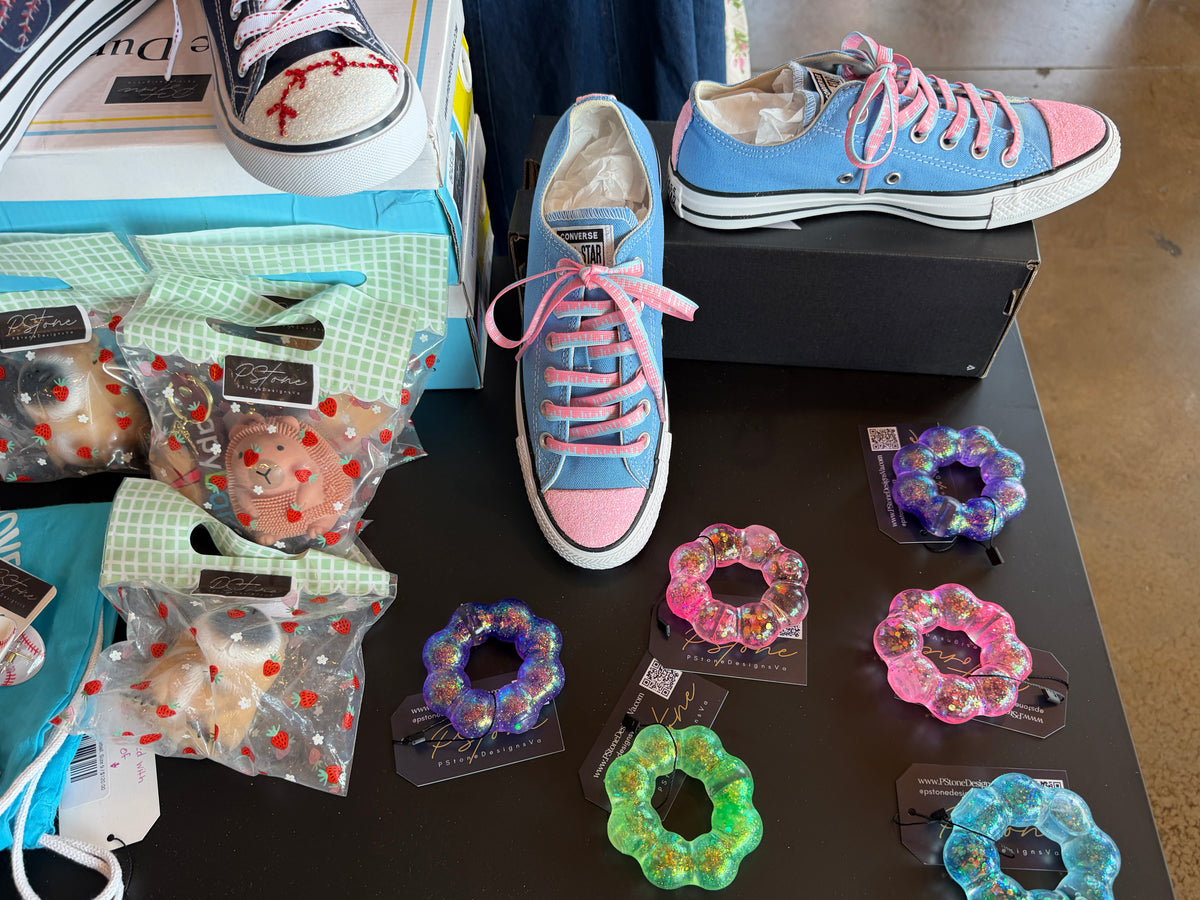 A pair of light blue and pink Converse-style low-top sneakers displayed on a black table, alongside several colorful squishy bracelets. A shoebox is visible behind the right shoe, and jeans appears at the edges.