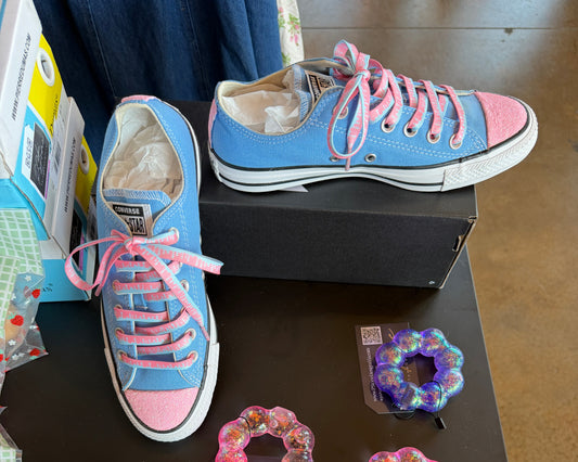 A pair of light blue and pink Converse-style low-top sneakers displayed on a black table, alongside several colorful squishy bracelets. A shoebox is visible behind the right shoe, and jeans appears at the edges.